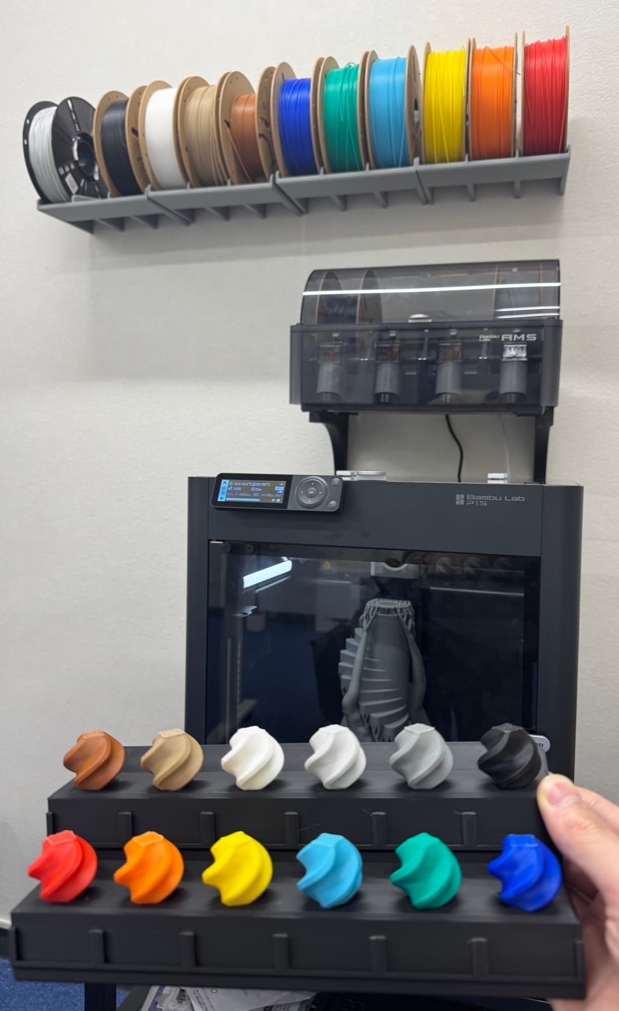 造形中の3Dプリンター。上の壁ラックには色とりどりのフィラメントが並び、手前には出力した色見本が置かれている